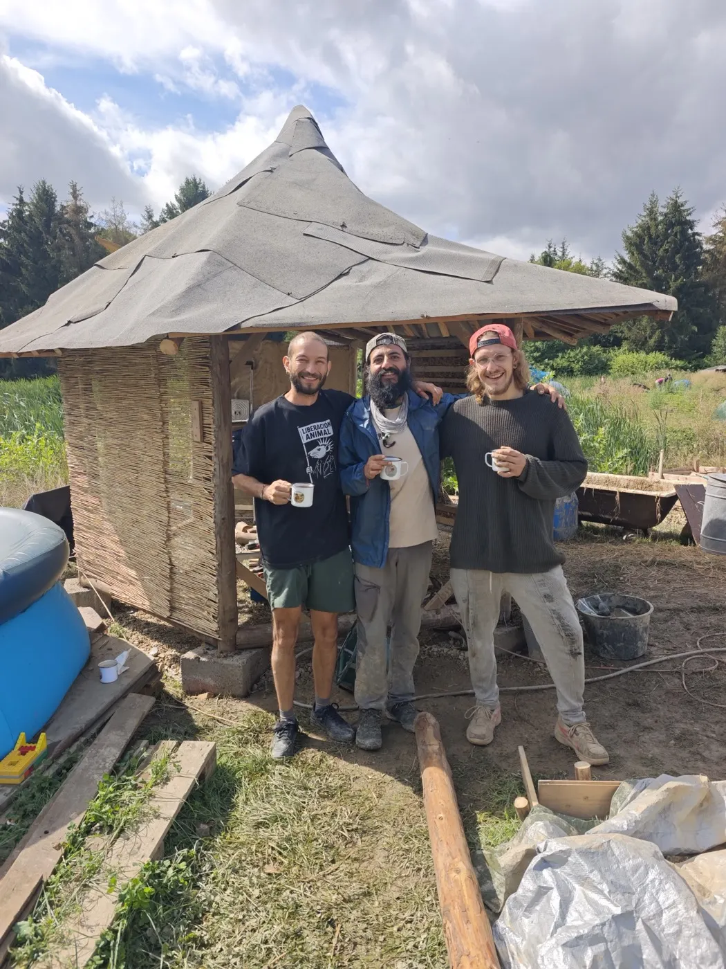 Drei Teilnehmende mit Kaffeebechern vor der Lehmhütte